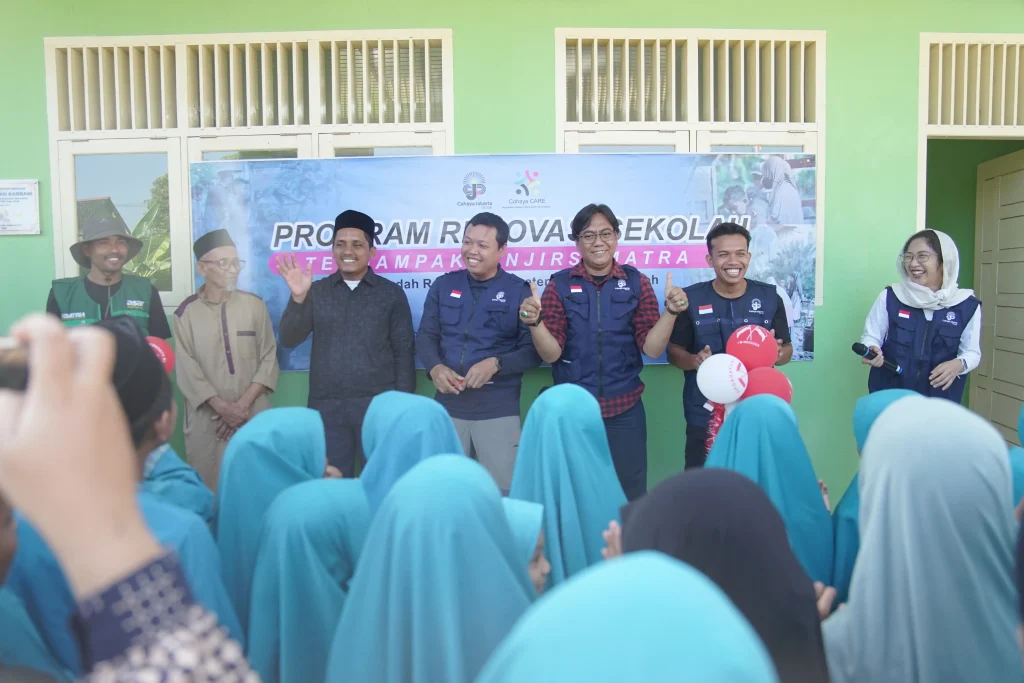 Group of organizers in blue vests posing and smiling in front of a banner, with girls in blue headscarves in the foreground.","People on stage at a community program, posing for photos behind balloons and a banner.","Five adults in blue vests and traditional attire celebrate a moment, raising hands as students watch.","Educational outreach event with staff and volunteers in blue vests, banner behind them.","Joyful scene of volunteers and students at a school program, turquoise hijabs in the crowd.