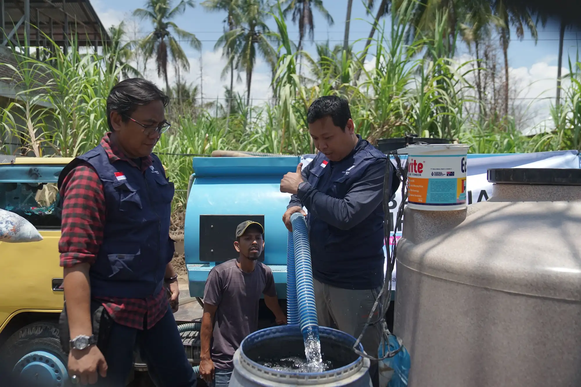 Three men in work vests operate blue industrial equipment outdoors, managing hoses and water flow into a barrel.